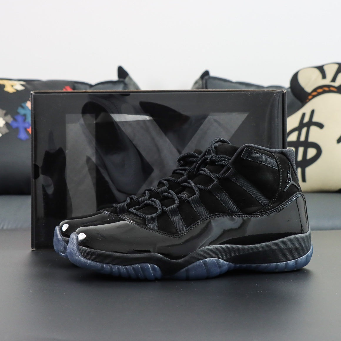 Air Jordan 11  Retro Cap and Gown. Black high-top sneakers with blue soles on a reflective surface, with a black box in the background.