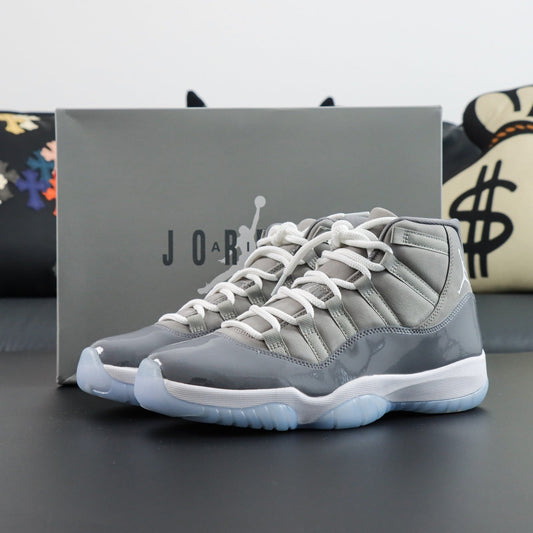 Air Jordan 11  Retro Cool Grey 2021. Sneakers with white laces and soles on a dark surface, with a Jordan box in the background.