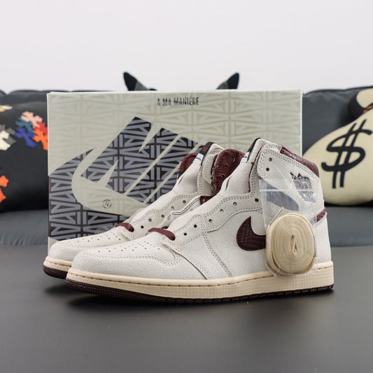 Air Jordan 1 x A Ma Maniére Retro OG High. White high-top sneakers with maroon accents on a dark surface, with a box in the background. 