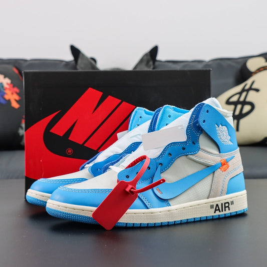 Blue and white sneakers with a red tag on a dark surface, with a Nike box in the background. Air Jordan 1 x Off-White Retro OG High University Blue
